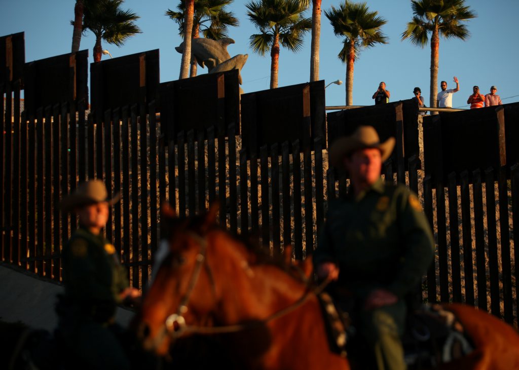 People in Mexico wave at U.S. Border Patrol agents on horseback patrolling the U.S.-Mexico border fence near San Diego, Ca...
