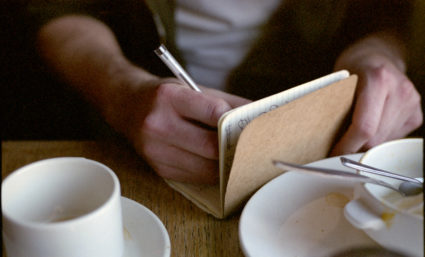 A writer sits in a cafe surrounded by teacups. He is writing in a notebook. The shot is a close up of his hands.. Photo by Getty creative