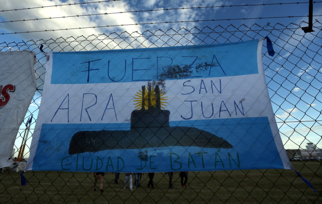 People walk behind an Argentine national flag displayed on a fence, in support of the 44 crew members of the ARA San Juan submarine who are missing at sea, at an Argentine naval base in Mar del Plata, Argentina, November 22, 2017. The words on the flag read: "ARA San Juan, be strong". REUTERS/Marcos Brindicci - RC1D573DDD00