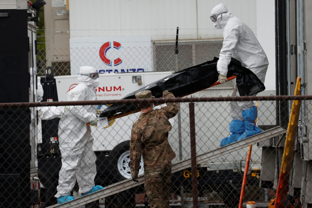 Workers wearing personal protective equipment (PPE) move the body of a deceased person from a refrigerated truck trailer s...
