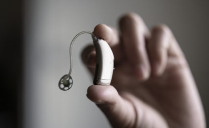 A white hand holds up a hearing aid