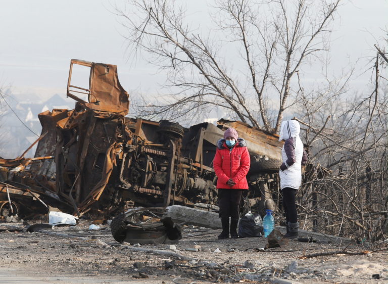 Local residents stand next to a destroyed vehicle in the besieged city of Mariupol