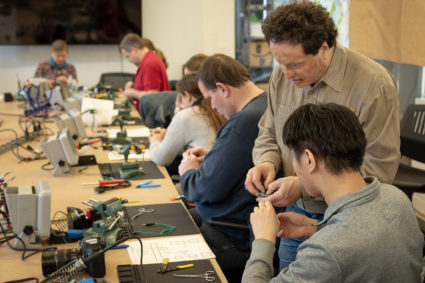 Amazon principal accessibility researcher Joshua Miele teaches workshop attendees how to solder. People sit at a long, large table holding tools in their hands, and Miele stands beside one attendee while explaining something to them.
