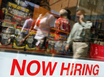 Pedestrians walk past a "Now Hiring" sign in the window of a GNC shop in Boston