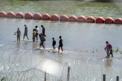 Migrants walk by a string of buoys placed on the water along the Rio Grande border with Mexico in Eagle Pass, Texas, on July 15, 2023, to prevent illegal immigration entry to the US. Photo by SUZANNE CORDEIRO/AFP via Getty Images