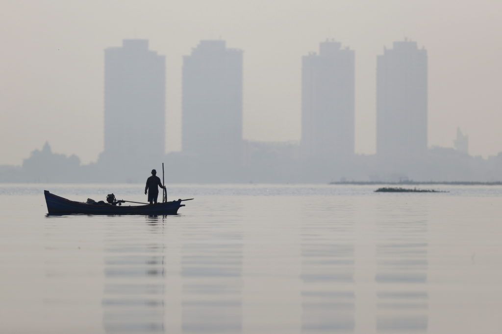 A fisherman stands on a wooden boat as smog covers high rise buildings at the north coast of Jakarta