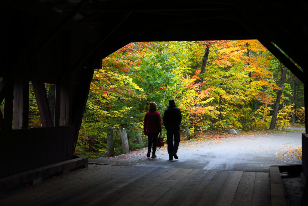 Couple leave covered bridge with trees in autumn