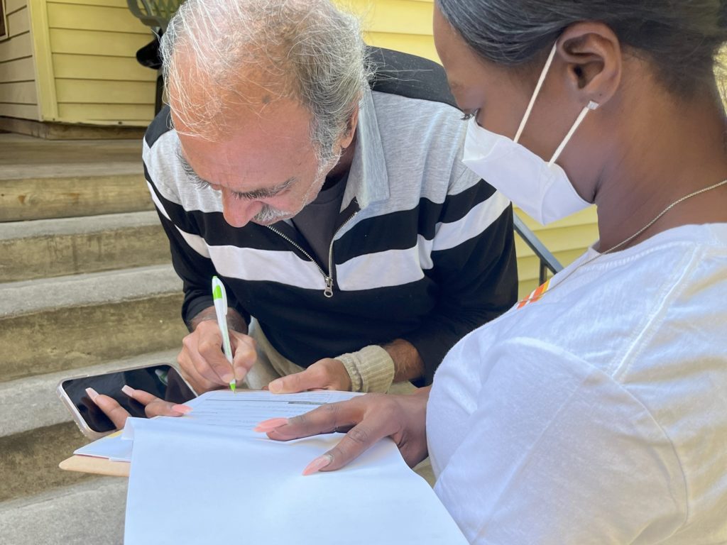 A Detroit Disability Power canvasser holds papers on a clipboard to register an elderly resident who is bent over holding a pen and filling out a form to vote in Detroit's Core City Neighborhood. Oct. 11, 2022. Photo courtesy of Rami Alvarez/Detroit Disability Power