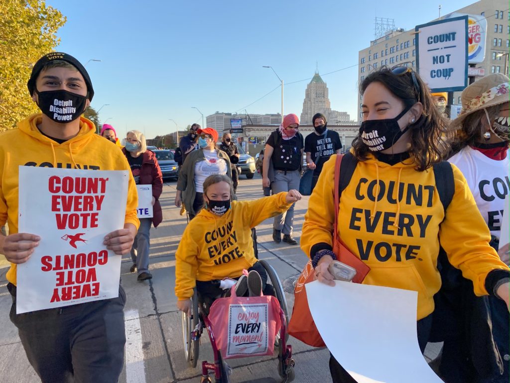 Activists from Detroit Disability Power march through Detroit on Election Day 2020 holding signs and wearing bright yellow hoodies that say Count Every Vote and masks that say Detroit Disability Power. A wheelchair user raises her fist in the air.
