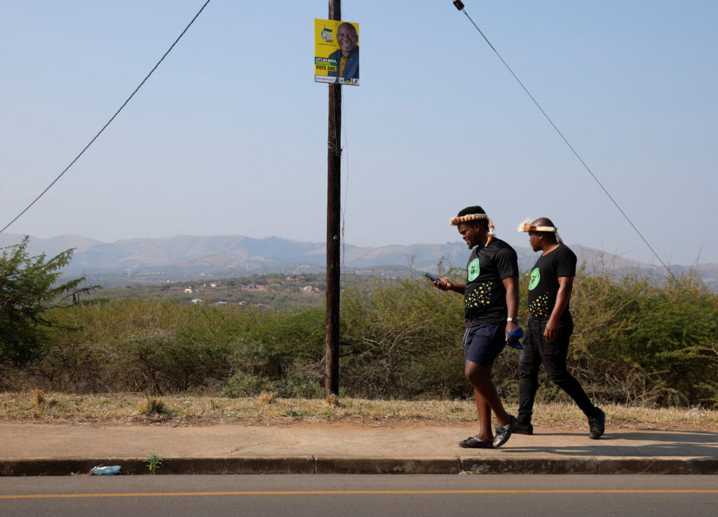 Men in uMkhonto weSizwe (MK) t-shirts walk past a poster of the African National Congress (ANC), in Nkandla