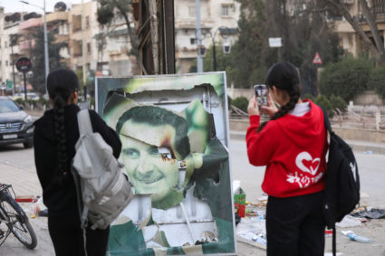 Women use their mobile phones near a damaged picture of Syrian President Bashar al-Assad as people celebrate, in Qamishli