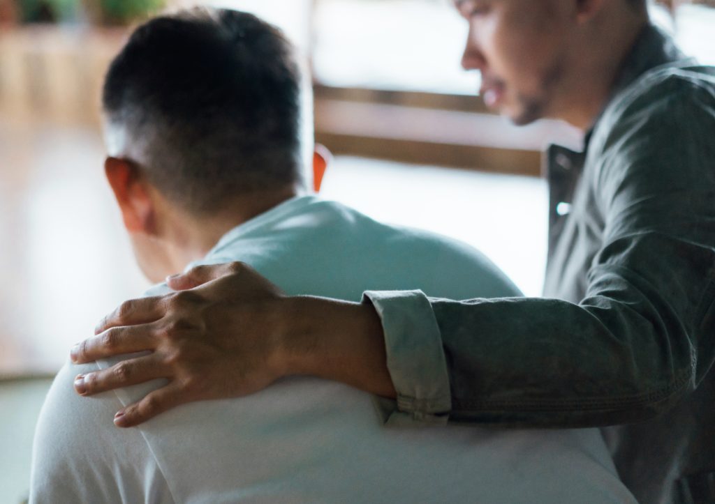 Rear view of son and elderly father sitting together at home. Son caring for his father, putting hand on his shoulder, com...