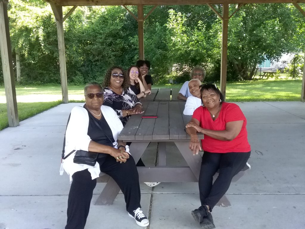 A group of people sitting at picnic tables in a park for a meeting to discuss Ypsi Farmers and Gardeners Oral History Project and the Generational Earn and Learn program at West Willow Community Garden