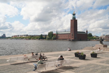 People rest on benches in the sunshine on the island of Riddarholmen with the City Hall in the background in Stockholm
