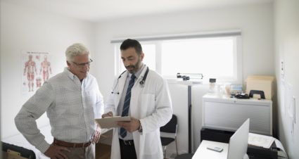 Male doctor with clipboard talking to senior patient in examination room