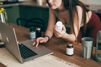 Close-up shot of young Asian woman holding a medicine bottle, reading from a laptop