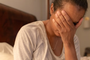 black woman holds head in hand sitting on bed