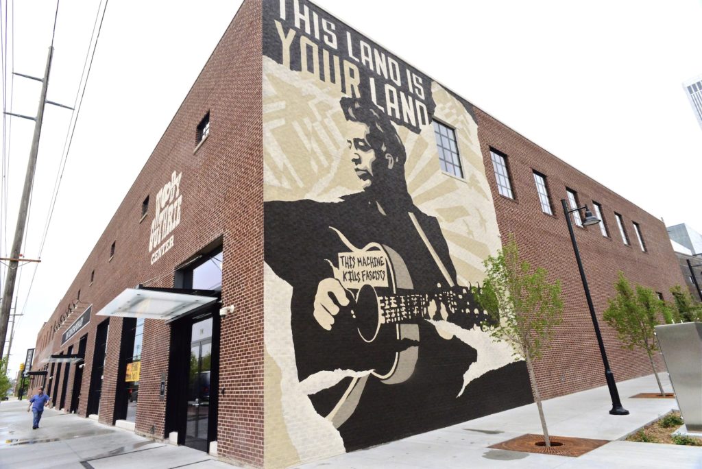 A mural of Woody Guthrie with the title of his most famous song emblazoned across the top guides visitors to the Woody Gut...