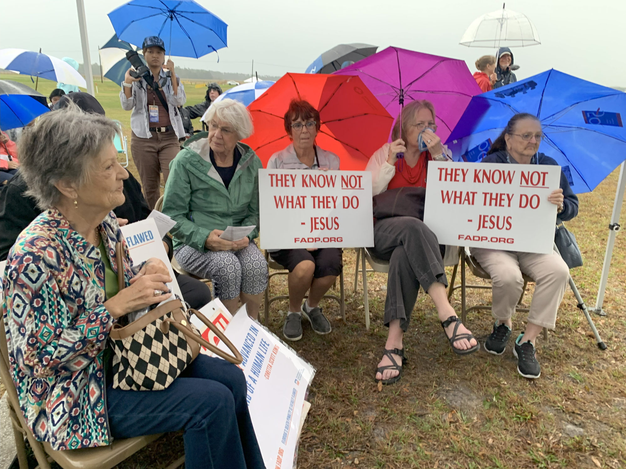 A row of seated demonstrators hold up umbrellas to shield them from the heat and signs that say in all caps They Know Not What They Do, which is attributed to Jesus Christ. This scene was captured on the day of Michael Bernard Bell’s execution on July 15.