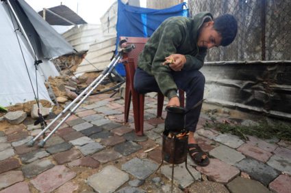 Displaced Palestinian Mahmoud Abu al-Kas, who has an amputated leg, sits tending a small fire in front of his family’s mak...