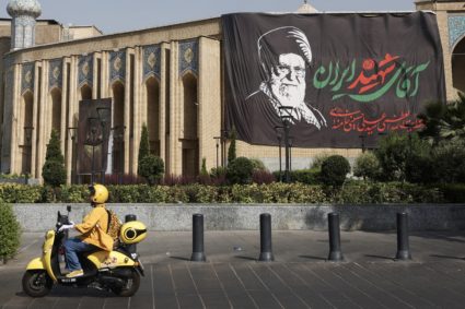 An Iranian woman rides a motorcycle next to a banner with a picture of the late Supreme Leader of Iran, Ayatollah Ali Kham...