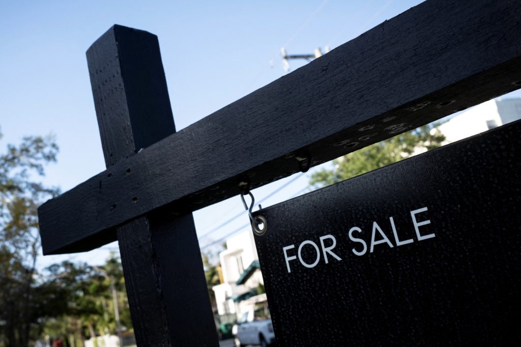 A 'For Sale' sign stands in front of a suburban home with autumn leaves on the ground