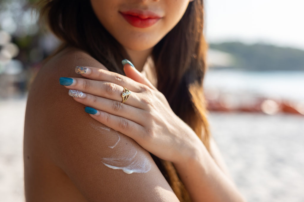 Beautiful happy cute woman applying suntan cream to her shoulder with beach background
